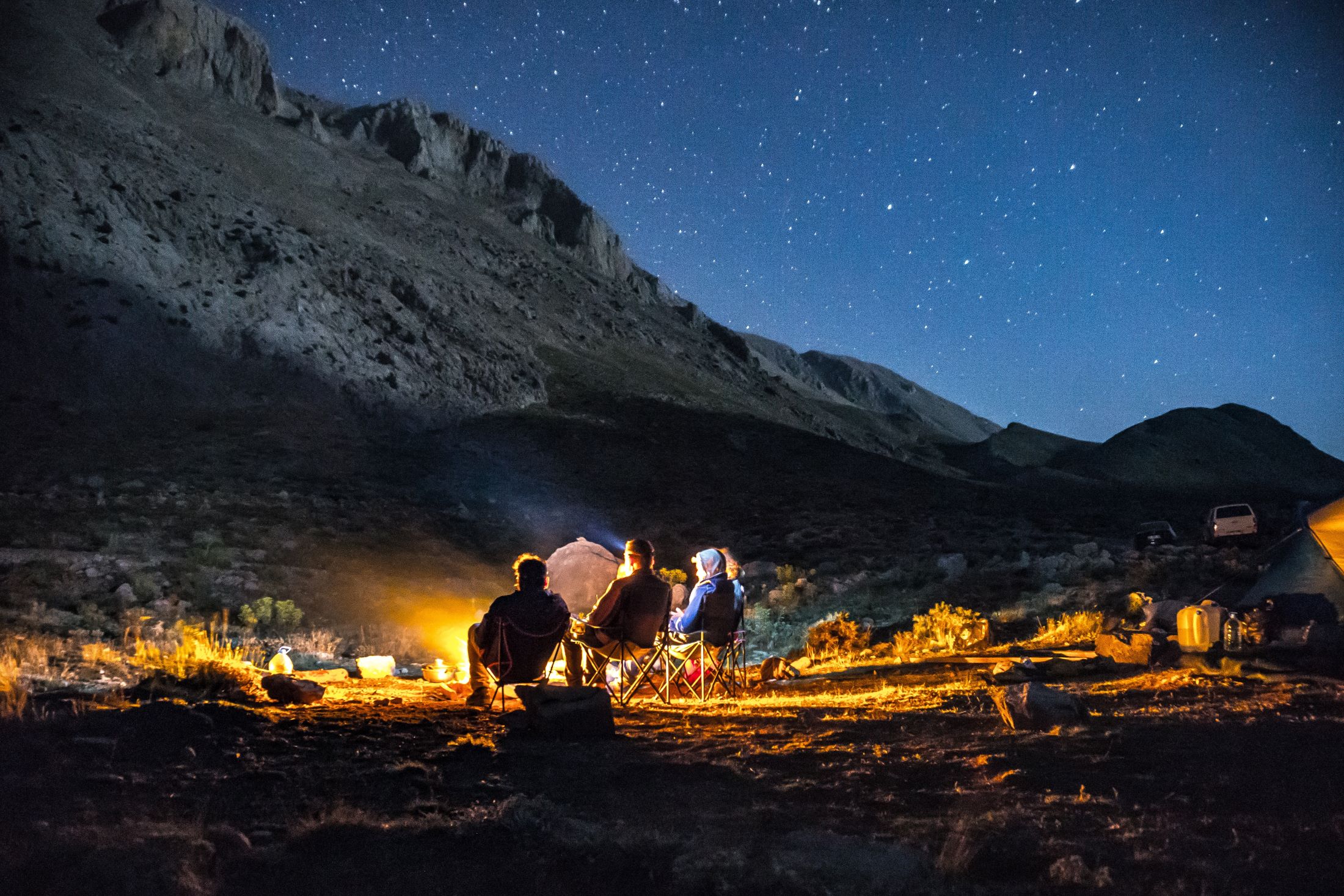 A group of campers at night sitting around a fire at the base of a mountain in the desert. 